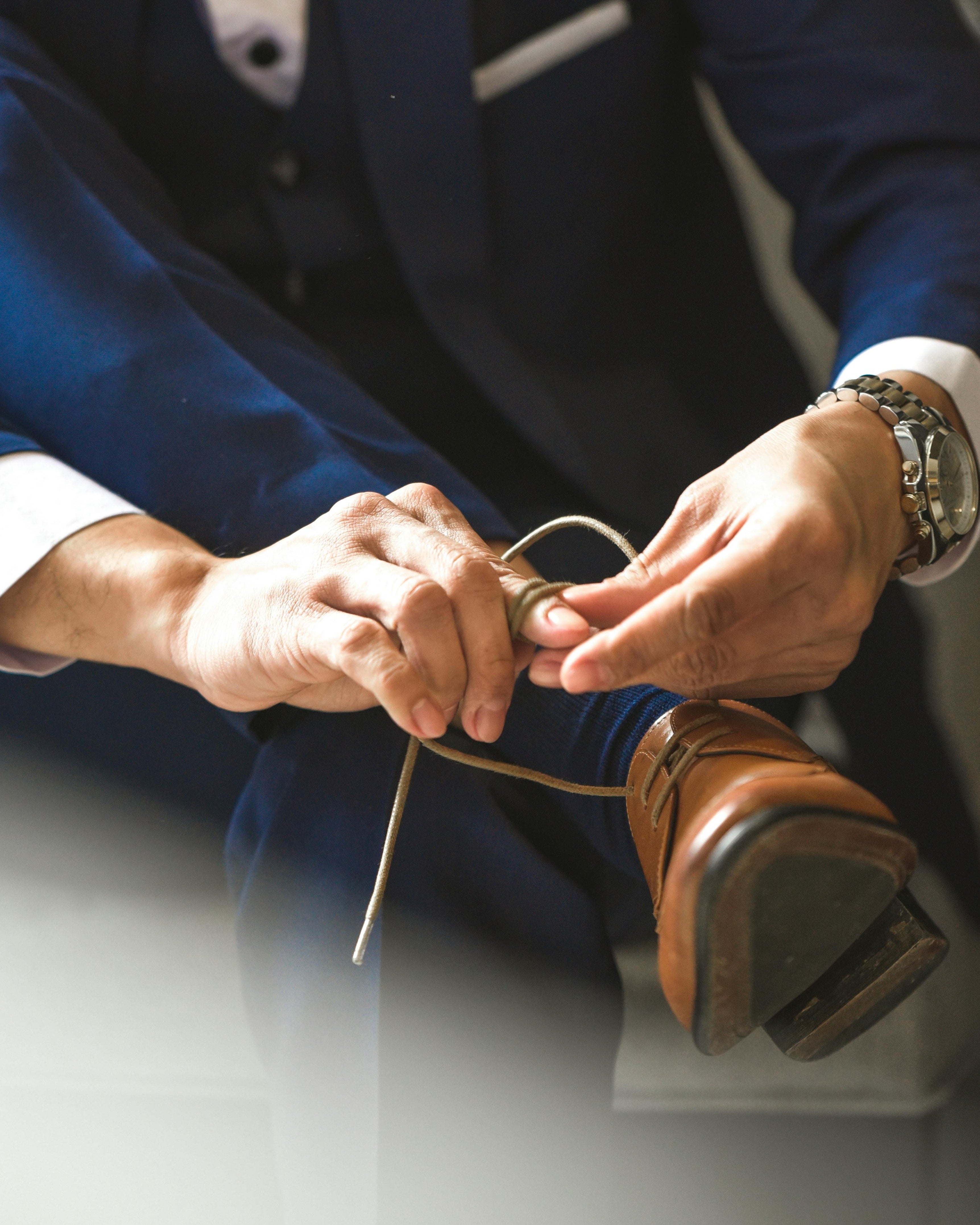 Man in a blue suit with a floral boutonniere tying his brown shoe.
